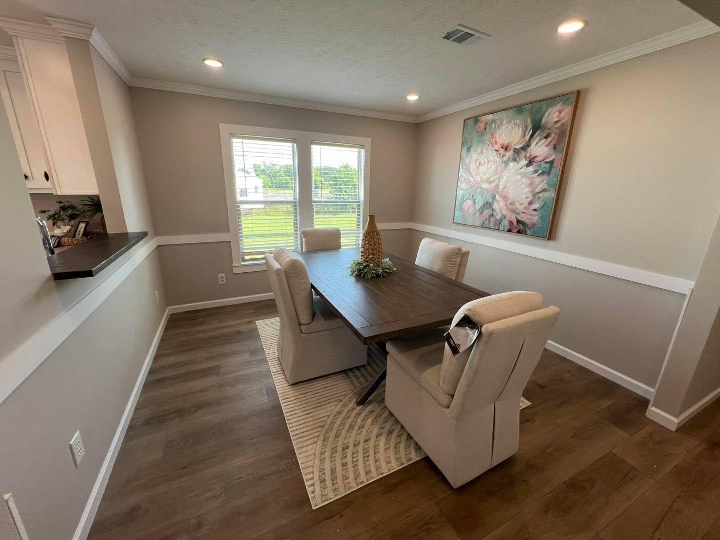 Modern luxury manufactured home dining room featuring a solid wood dining table for four with upholstered beige chairs, a large floral canvas art piece, and a stylish patterned area rug over high-end wood-look vinyl flooring, all illuminated by recessed lighting and natural light from twin windows for a sophisticated 4-bedroom move-in ready turn-key residential design.