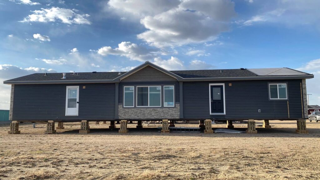 Modern modular home under construction on concrete block piers in open prairie landscape, dark gray vinyl siding with stone accent base, gable roof, large windows, navy blue front door, spacious single-story manufactured bungalow exterior view against blue sky with scattered clouds