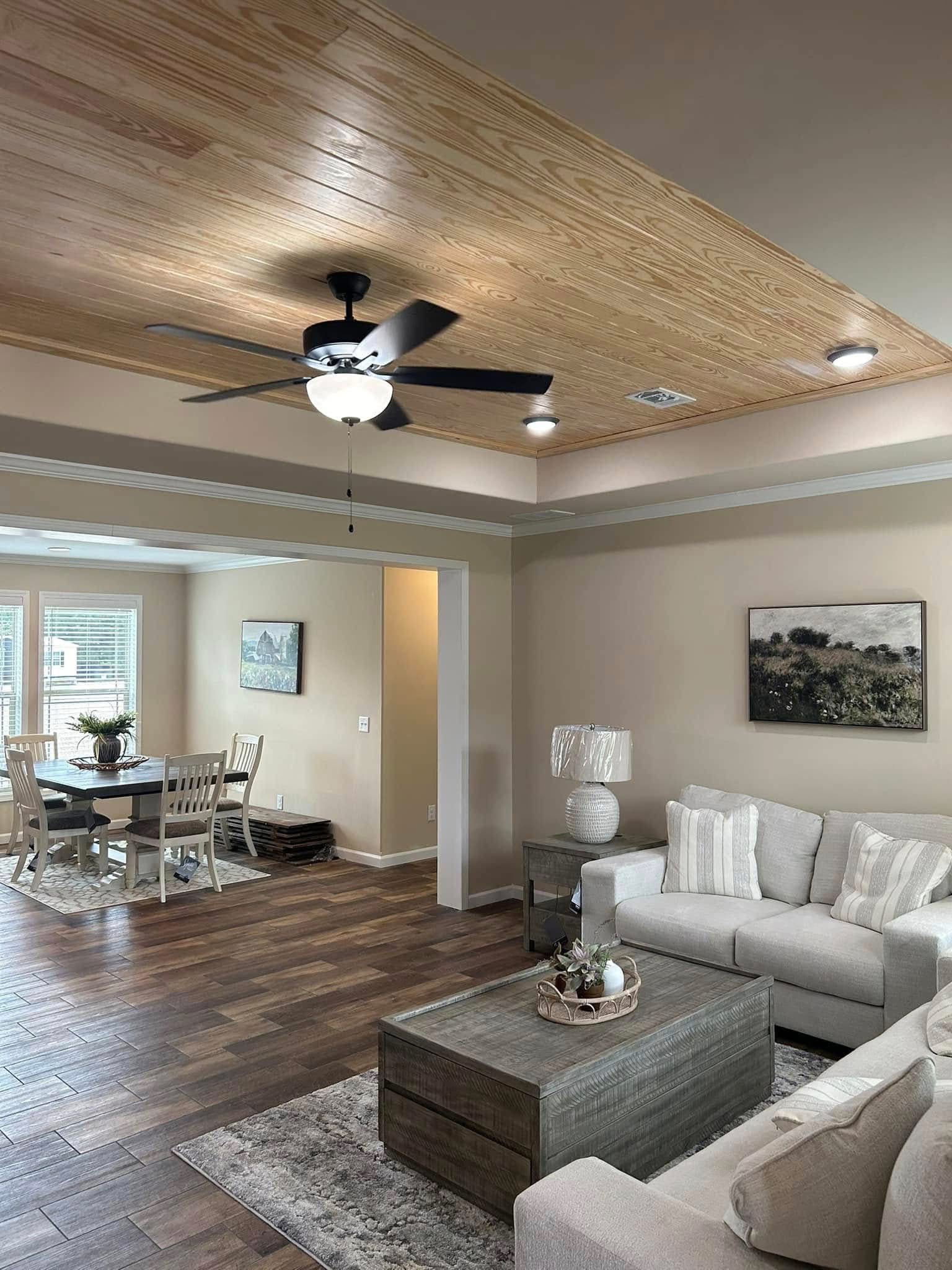 Spacious living and dining area with a warm, wood-paneled ceiling featuring a black ceiling fan and recessed lighting, complemented by beige walls, dark wood plank flooring, a comfortable sectional sofa with striped throw pillows, a rustic wooden coffee table, and a dining set with chairs near a window.