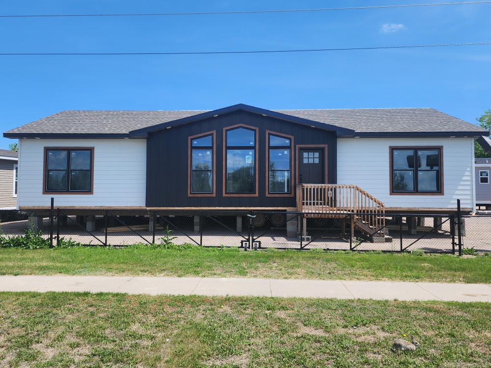 A modern, white modular home with contrasting dark brown accents and large windows. A small wooden staircase leads to the entrance. Bright, clear sky.