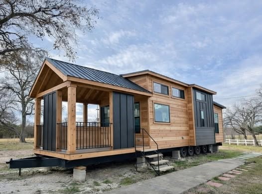 Modern luxury tiny home on wheels featuring a sophisticated combination of natural cedar wood siding and black vertical metal panels, a durable black metal roof, and an integrated covered front porch with timber pillars and industrial black railings. This high-end mobile architectural design showcases large energy-efficient windows, a triple-axle trailer chassis for portability, and a contemporary rustic aesthetic perfect for off-grid living or a premium backyard guest house.