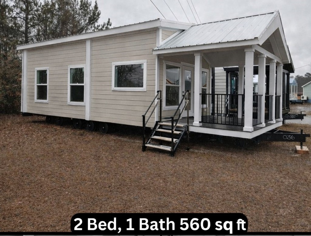 Exterior view of a light tan modular tiny home with a white metal roof, a covered porch with white columns and black railings, and dual-pane windows, situated on a gravel lot with visible trailer chassis and wheels, indicating it is a mobile or manufactured dwelling, with text overlaying the bottom reading "2 Bed, 1 Bath 560 sq ft".