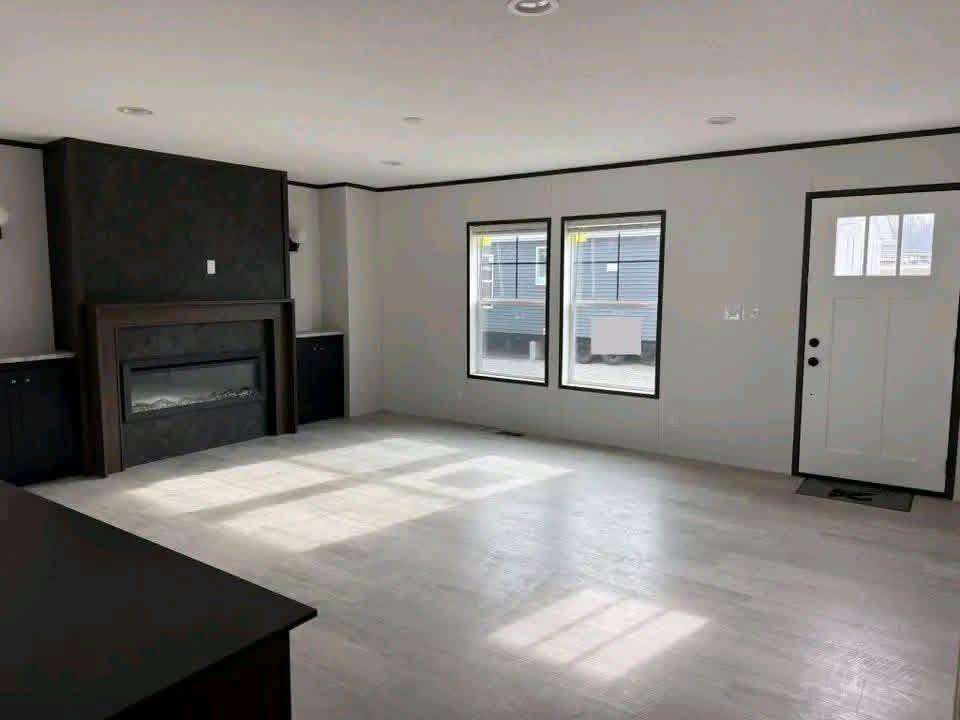 Modern luxury manufactured home living room featuring a minimalist black electric fireplace as the focal point, complemented by bright white walls, large black-trimmed energy-efficient windows that flood the space with natural light, and high-end grey wood-look vinyl flooring for a clean and contemporary move-in ready residential interior design.