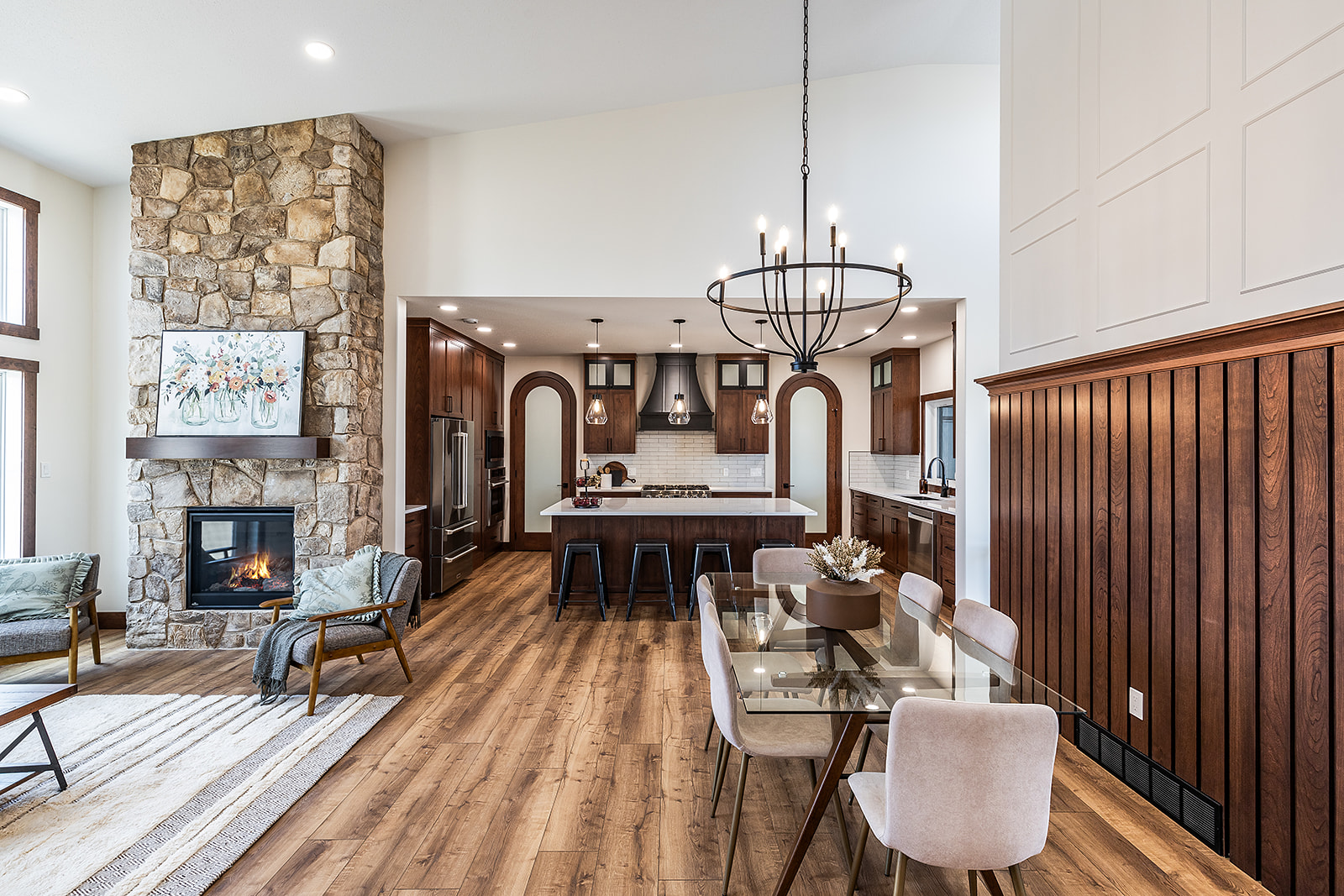 Spacious open-concept room with stone fireplace, modern kitchen, wooden floors, and elegant dining area. Warm tones create a cozy, inviting ambience.