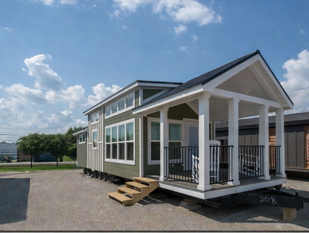 A modern tiny house with green siding and white trim, features large windows and a small front porch. It's set on a sunny day with a blue sky and clouds.