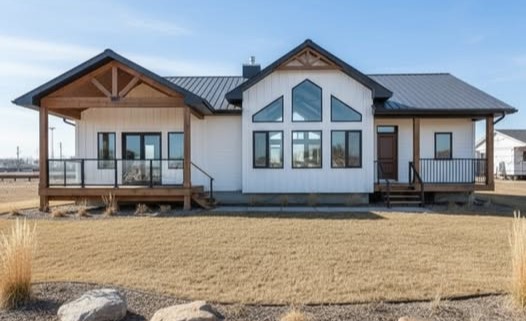 Modern white farmhouse-style modular home featuring a black metal roof, natural wood timber gables, and a spacious covered front porch with a black railing. The exterior architectural design showcases a stunning central peak with large geometric windows, including a prominent arched window, framed in black to create a contemporary aesthetic. This luxury prefab residence is situated on a landscaped lot with dry grass and decorative boulders, highlighting a perfect blend of rustic charm and modern industrial elements.