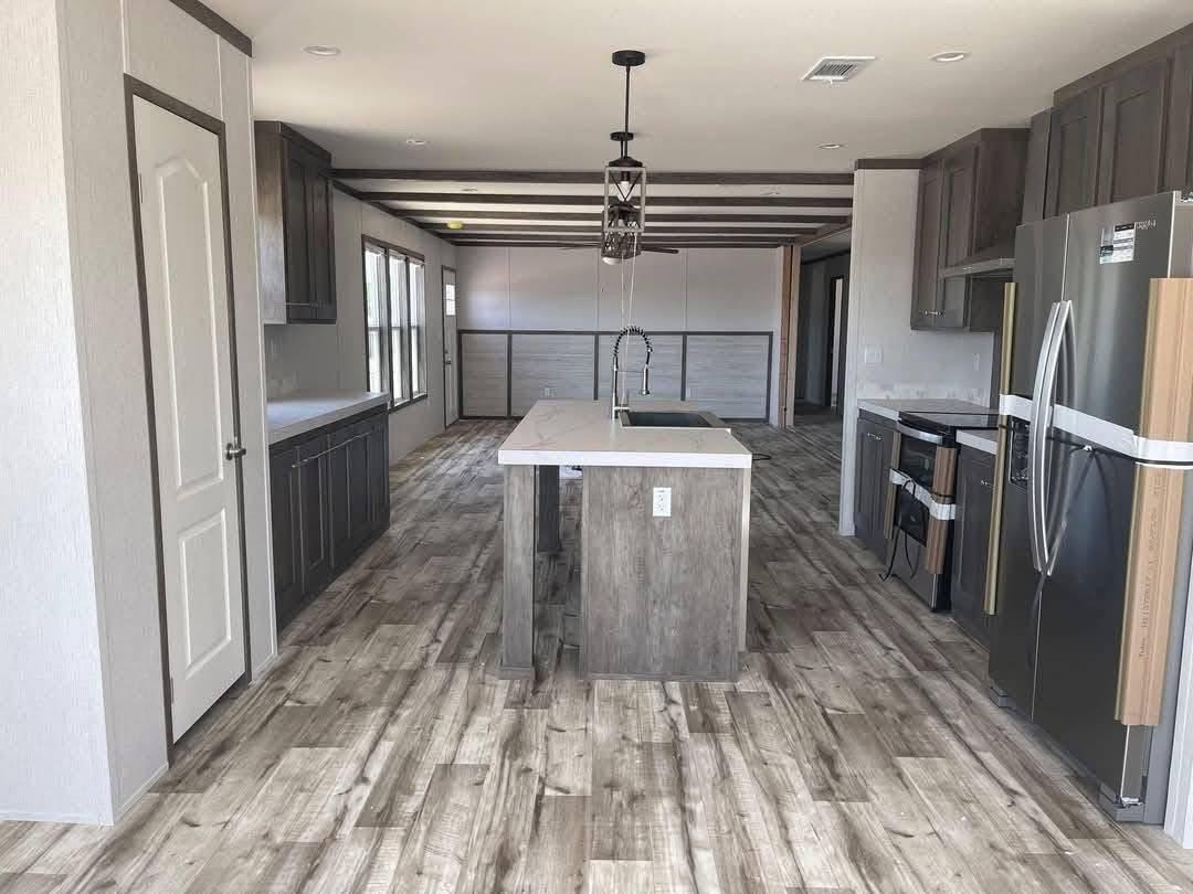 Spacious open-concept modern kitchen and living area in luxury manufactured home featuring large central island with white quartz countertop and farmhouse sink, dark gray shaker cabinets, stainless steel appliances including double-door refrigerator and range, rustic wood-look luxury vinyl plank flooring, exposed dark beam ceiling with black industrial pendant lights, beige accent walls, recessed lighting, ceiling fan, large windows for natural light, and open layout perfect for family gatherings – ideal for high-end modular home real estate listings and affordable luxury living.