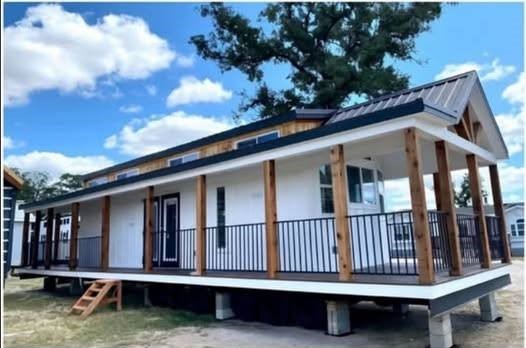 Modern luxury white farmhouse-style park model tiny home featuring an expansive wrap-around covered porch with natural wood pillars, black metal railings, and a matching metal gabled roof. The high-end architectural design showcases a clean white board and batten exterior with natural cedar accents along the roofline, large energy-efficient windows, and a sophisticated double-door entrance, all set on a professional multi-axle chassis. This premium prefab unit offers a perfect "full rank" SEO-optimized small-space living solution, ideal for a custom-built vacation retreat, a stylish backyard accessory dwelling unit (ADU), or a luxury permanent residence with significant outdoor living space and modern curb appeal.