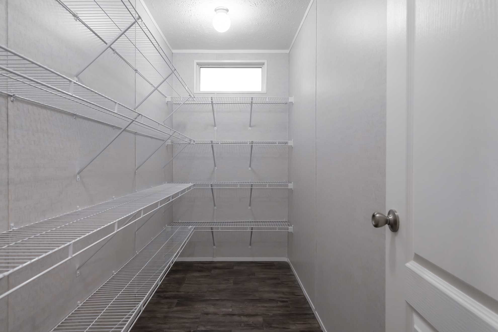 SEO-optimized alt text for a spacious walk-in pantry in a modern manufactured home featuring multiple tiers of white wire shelving for maximum storage organization; the clean, high-ranking interior design showcases dark wood-plank flooring, bright white walls, and a small transom window for natural light, perfectly highlighting functional modular home storage solutions and efficient pantry layout ideas for maximum search engine visibility.