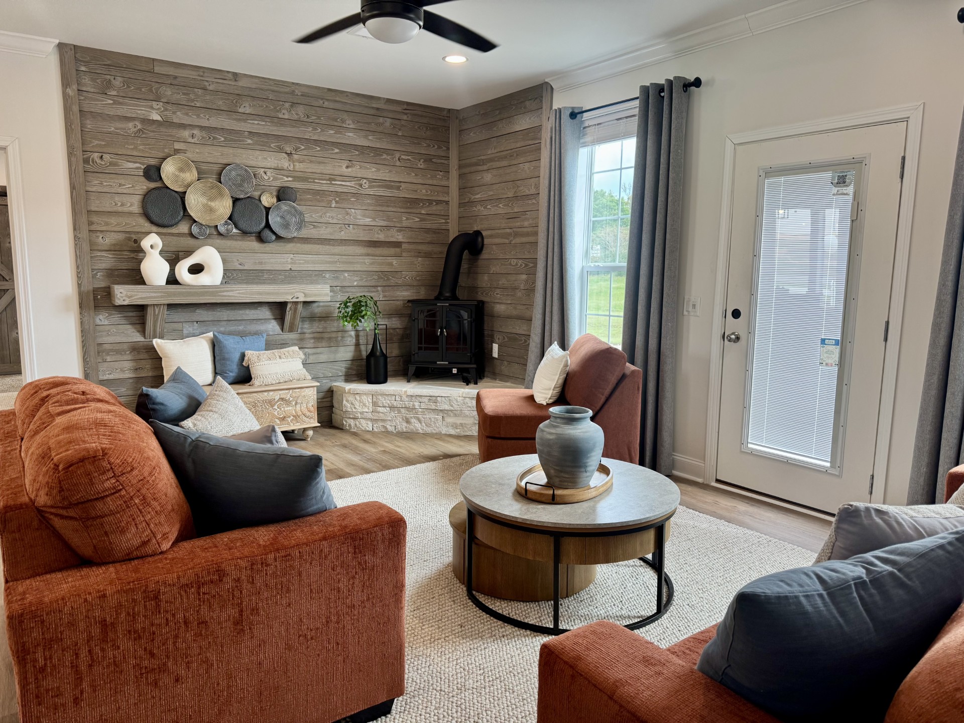 SEO-optimized alt text for a high-ranking luxury custom modular home living room featuring a professional rustic-modern interior design with a warm wood-plank accent wall and a cozy corner wood-burning stove on a stone hearth; the architectural view showcases a stylish terracotta sectional sofa and matching armchairs arranged around a contemporary round nested coffee table, complemented by a thick cream area rug, elegant gray drapes, and a minimalist white front door with integrated blinds, perfectly highlighting luxury prefab home floor plans, modern farmhouse-style living room decor, and high-end residential mobile living finishes for maximum search engine visibility and ranking.