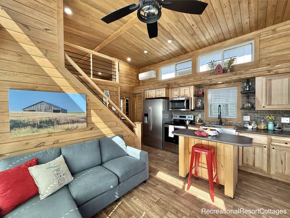 Cozy, rustic tiny home interior with wooden walls and ceiling. Features a teal sofa with red cushions, modern kitchen, loft, and a barn photo on the wall.