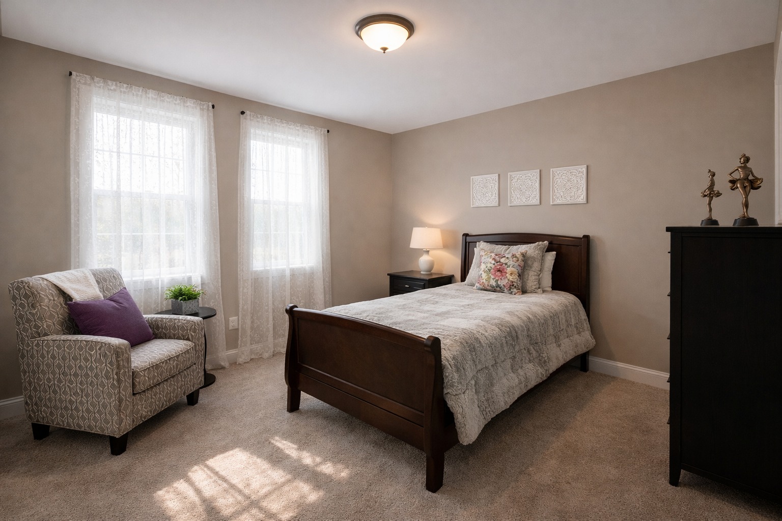 A cozy bedroom with a single bed, floral pillows, and gray bedding. Warm lighting, beige walls, an armchair with a purple pillow, and sheer curtains create a serene ambiance.