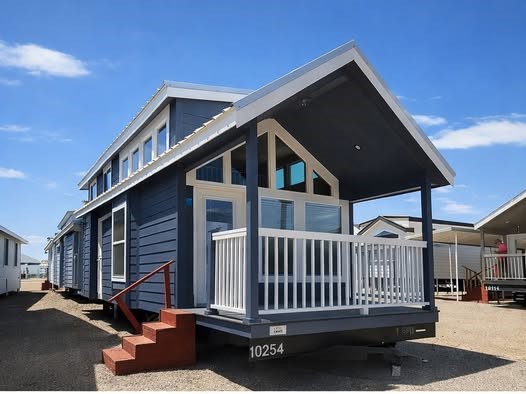 Blue tiny house with white trim and a covered porch, surrounded by gravel. It features large windows and red brick steps, set against a clear blue sky.