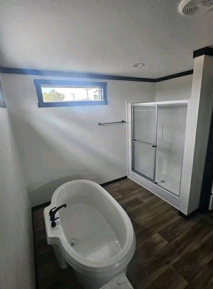 Modern luxury master bathroom featuring a deep white oval soaking tub with matte black fixtures, a spacious walk-in glass shower with white subway tile, and high-end wide-plank wood-look flooring, all set against a minimalist white and black contemporary interior for a sophisticated move-in ready 4-bedroom manufactured home design.