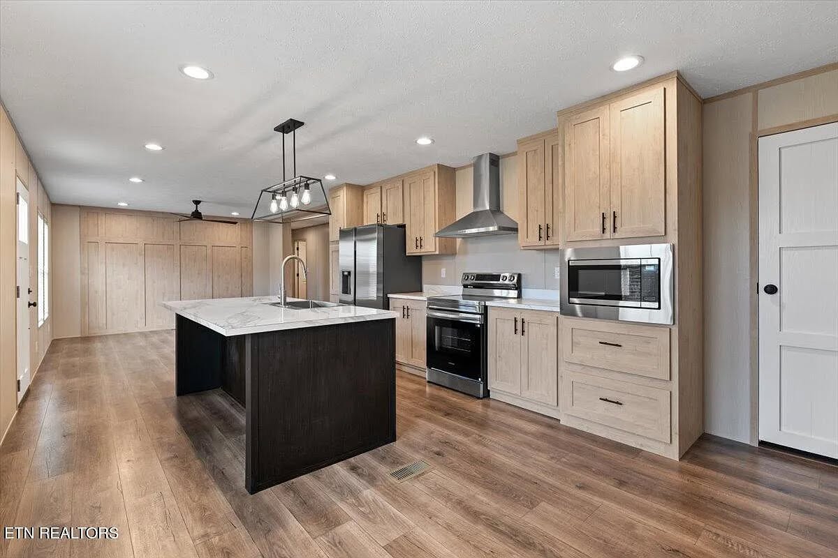 Spacious open-concept gourmet kitchen in modern modular manufactured home featuring large central island with dark charcoal black base and gleaming white marble-look quartz countertop with integrated undermount sink and brushed nickel faucet, light natural oak shaker-style cabinetry with black hardware extending to full-height pantry walls, built-in stainless steel appliances including side-by-side refrigerator double oven range microwave and dishwasher, powerful stainless range hood with contrasting backsplash, recessed ceiling lights pendant island lighting and ceiling fan, wide-plank dark walnut luxury vinyl flooring, neutral beige walls with crown molding, bright airy transitional farmhouse-style kitchen interior view in new single-story ranch bungalow