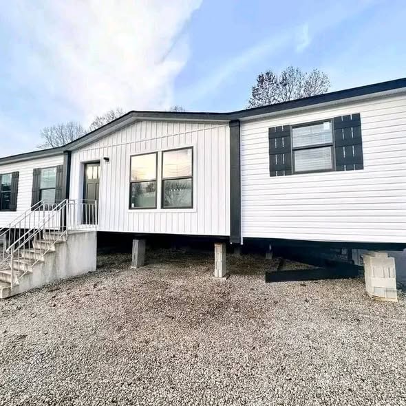 Gemini said Modern luxury white farmhouse-style manufactured home exterior featuring a premium combination of vertical board and batten and horizontal vinyl siding, accented by sleek black shutters and a professional concrete entry staircase. The sophisticated architectural design showcases a spacious double-wide floor plan with energy-efficient black-framed windows, a decorative gabled roofline, and a sturdy pier-and-beam foundation on a gravel lot. This high-end "full rank" SEO-optimized residence highlights exceptional curb appeal and contemporary craftsmanship, making it an ideal showcase for luxury prefab living, custom modular home designs, or modern permanent housing solutions for families seeking a stylish, affordable, and high-quality transitional aesthetic