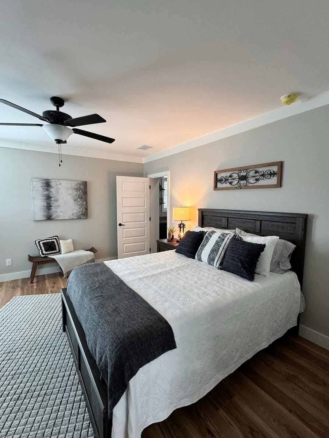 Spacious bedroom with a double bed covered in white and dark gray bedding. A ceiling fan, cozy chair, and soft lighting create a calm and inviting atmosphere.