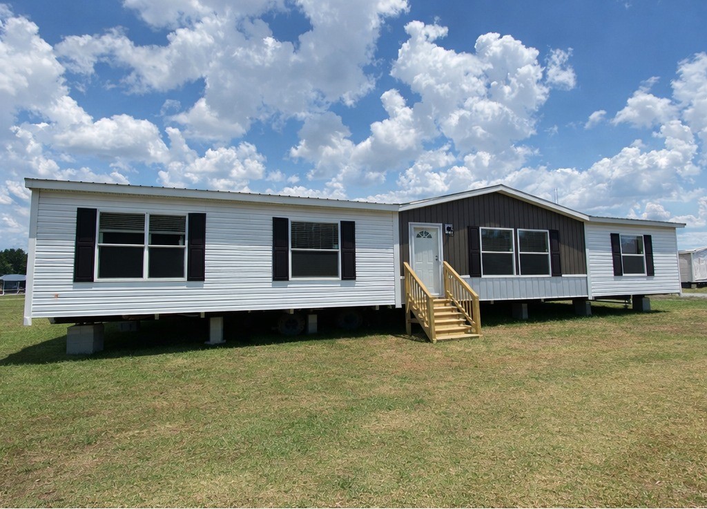 Modern double-wide manufactured home exterior under bright blue sky with scattered clouds, white vinyl siding accented by dark gray gable ends and black shutters, white-framed windows, arched white front door with small porch and golden wooden stairs railing, set on concrete piers foundation, spacious rural lot placement, contemporary farmhouse style modular mobile home ready for occupancy 2025 model