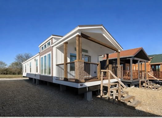 A modern tiny house with white siding and a small wooden porch sits elevated on concrete supports. Clear blue sky and trees in the background evoke a peaceful, minimalist vibe.