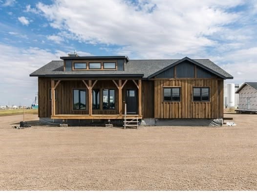 Rustic wooden house with a dark roof and large windows, set on a barren, gravelly landscape under a partly cloudy blue sky, conveying simplicity and solitude.