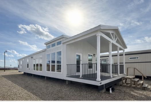 Modern white modular home with large windows and covered porch, set on gravel under a bright sun in a clear blue sky. Inviting and tranquil atmosphere.