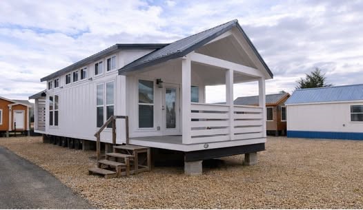 Modern tiny house on piers with white board and batten siding, black metal gable roof, spacious wrap-around front porch with white railings, large energy-efficient windows, wooden entry steps and deck, gravel foundation pad, charming farmhouse exterior design in a tiny home community – perfect for off-grid living, vacation cabin, or minimalist tiny house inspiration.