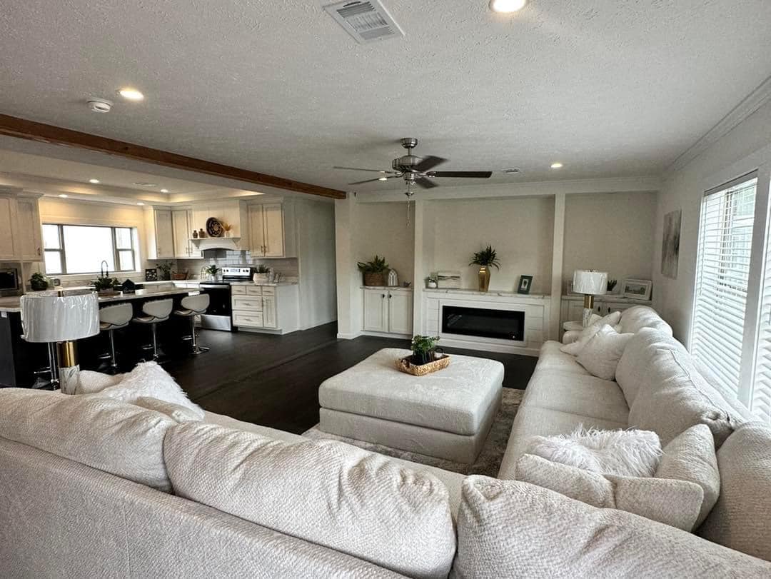 Spacious, modern living room and kitchen. Large white sectional sofa, central ottoman, cozy fireplace, ceiling fan, and bright decor create an inviting atmosphere.