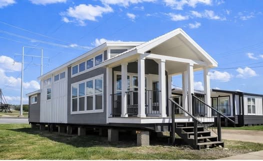 Modern tiny house on stilts with gray siding, large windows, and a white-columned porch. Set against a blue sky with scattered clouds, conveying a cheerful mood.