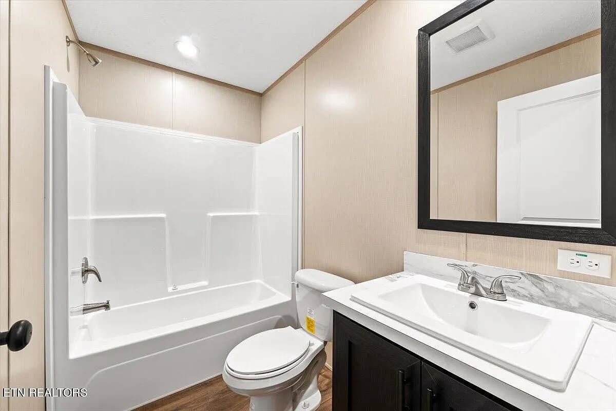 Modern compact bathroom in contemporary manufactured home featuring white tub-shower combo with built-in shelving, beige textured wall panels, dark wood vanity cabinet with white marble-look countertop, rectangular vessel sink, large black-framed mirror, brushed nickel faucets, recessed ceiling lighting, textured popcorn ceiling, and vinyl plank flooring – ideal for real estate photos of affordable modular homes.