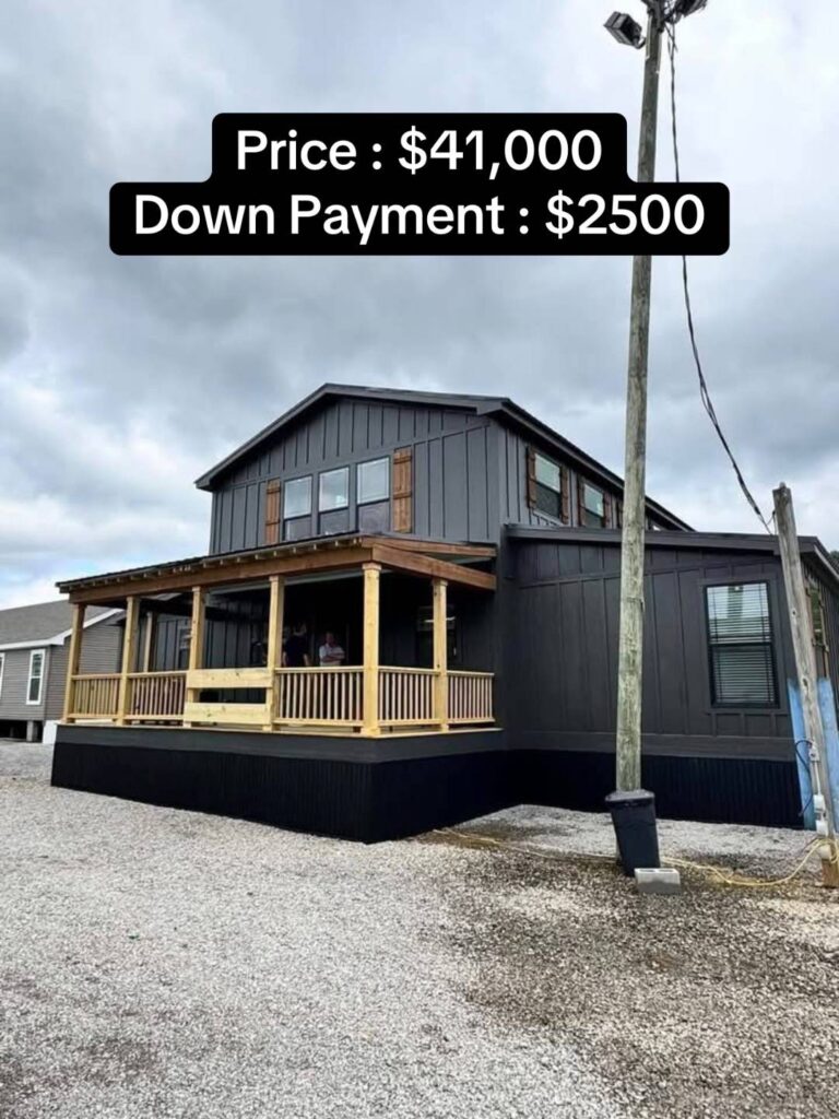 Two-story dark gray house with wood accents and covered porch. Price: $41,000, Down Payment: $2,500. Overcast sky and gravel driveway.