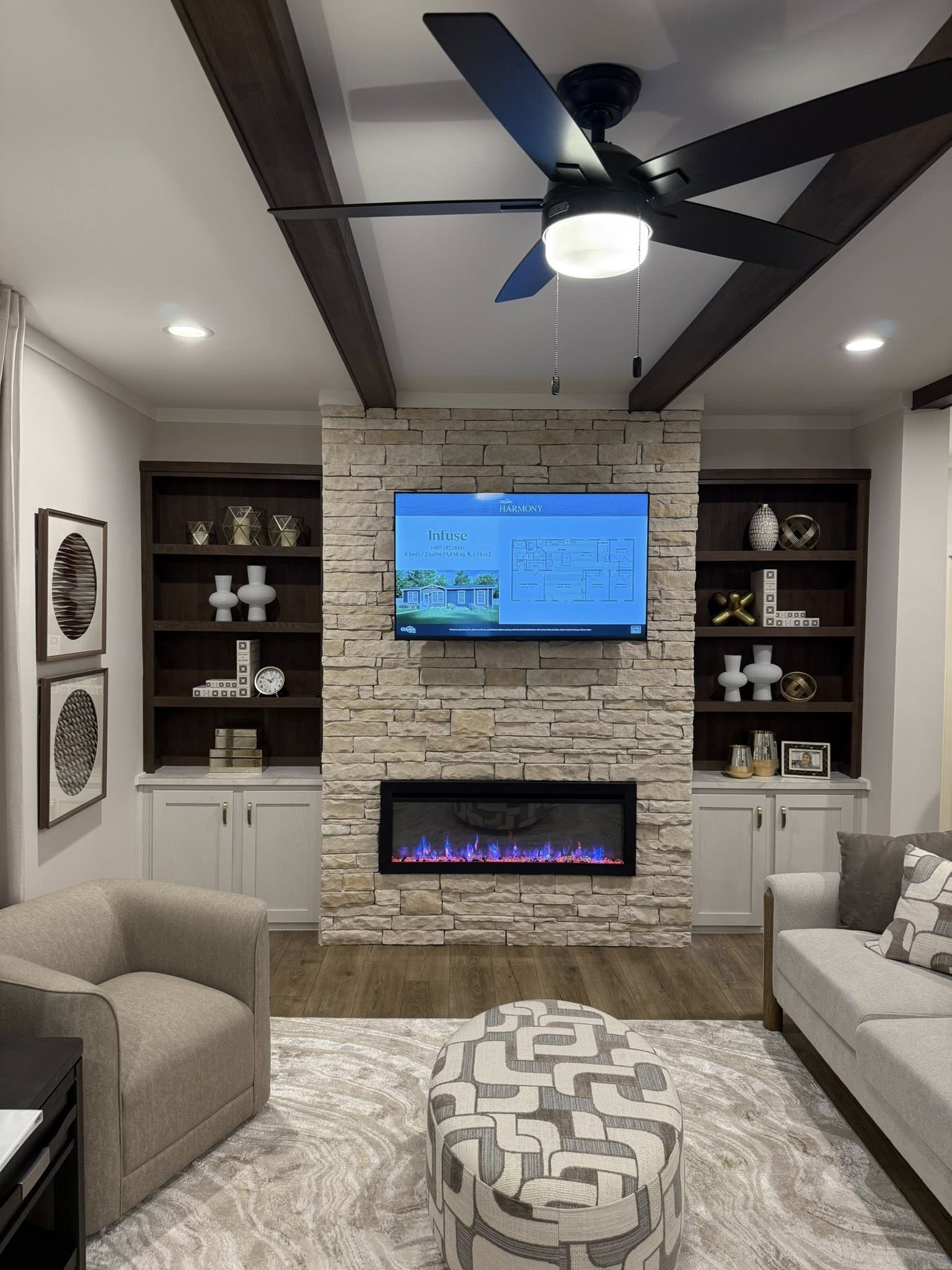 Living room with stone fireplace, mounted TV, and modern fan. Cozy seating, abstract ottoman, and shelves with decor create a warm, inviting space.