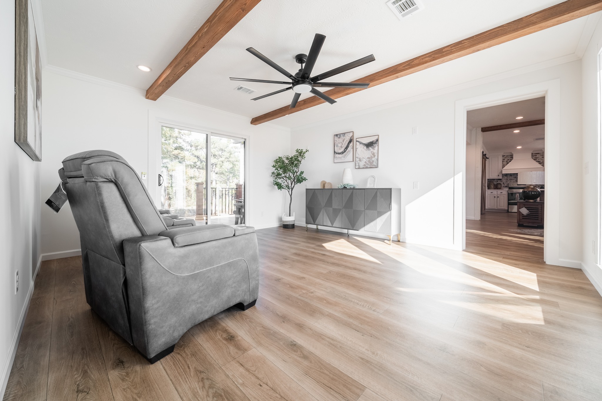 Modern living room with wooden floor, gray recliner on the left, ceiling fan, and wood beams. Sunlit space features wall art, sideboard, and potted plant.