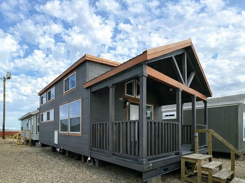 A modern tiny house with dark gray siding and a front porch stands against a backdrop of a cloudy blue sky, evoking a sense of coziness and simplicity.