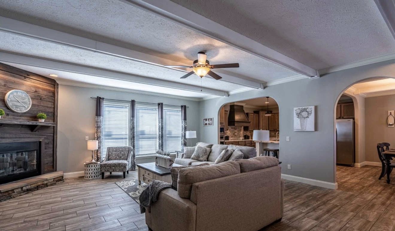 Spacious living room with gray walls, a large beige sectional sofa, and a wooden coffee table. A fireplace is on the left, and a ceiling fan above. Cozy and inviting.