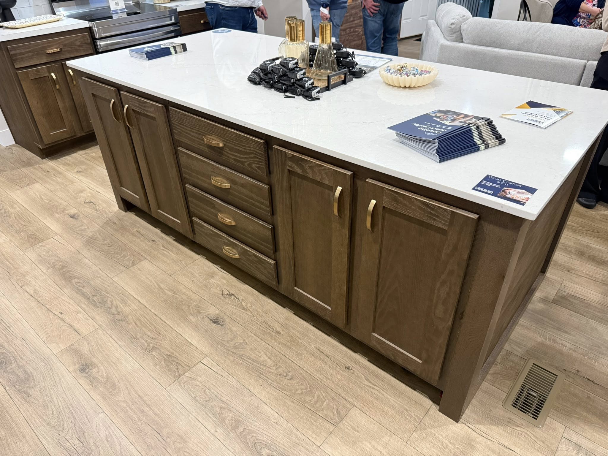 A kitchen island with a white countertop and dark wood cabinets, displaying brochures, gold bottles, and small black items, set on light wood flooring.