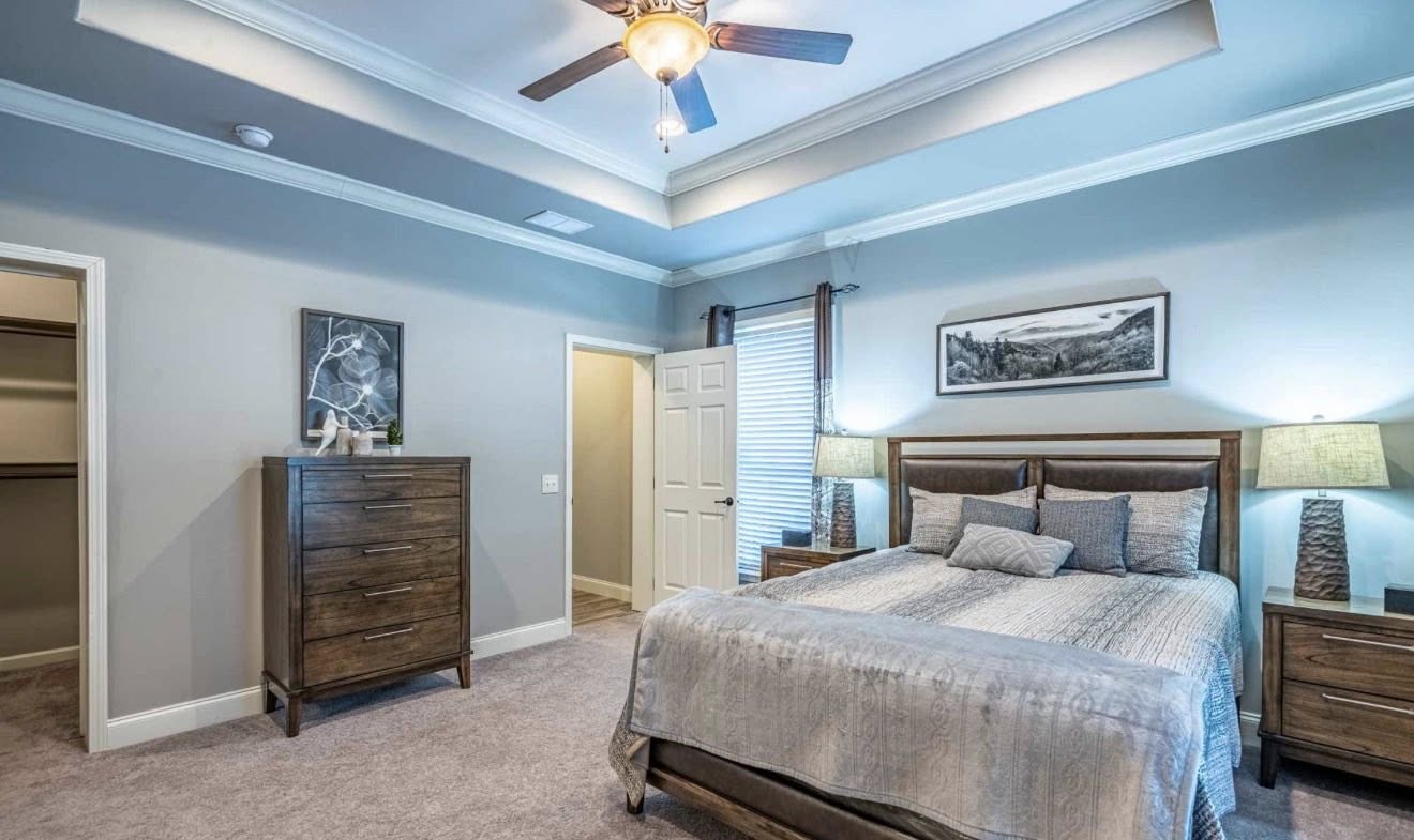A cozy bedroom with soft gray walls and carpet, features a wooden bed with gray bedding, matching nightstands, a dresser, and ceiling fan. Warm, inviting atmosphere.