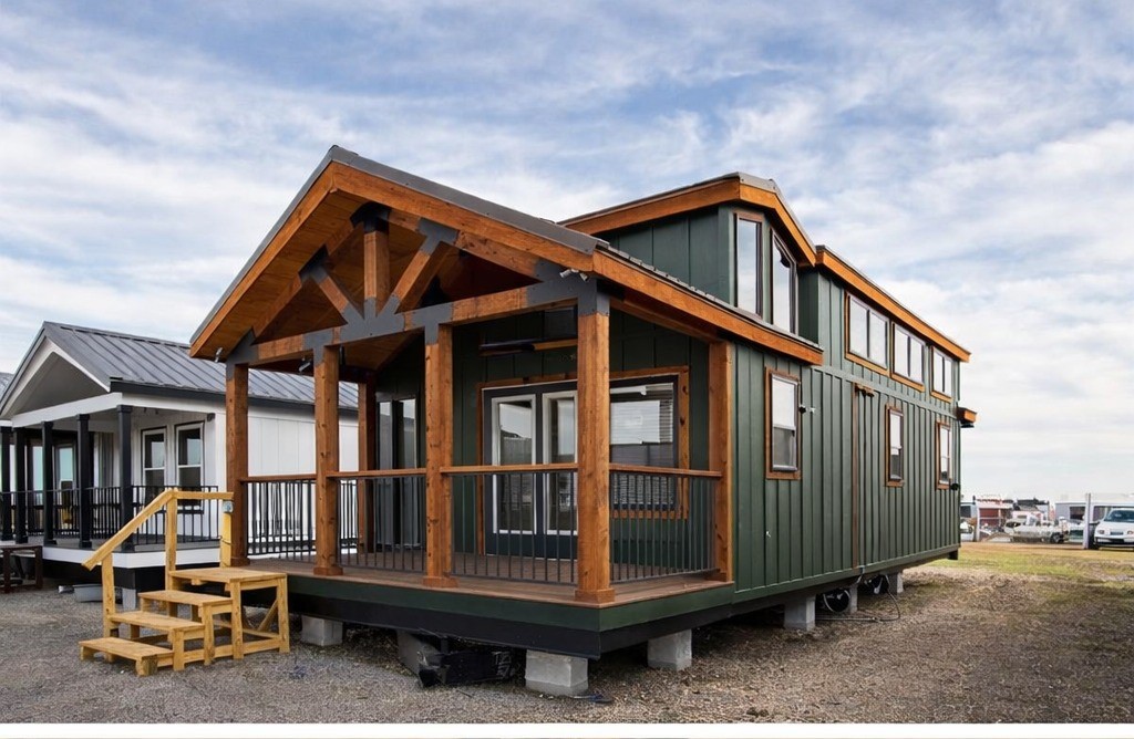 A green tiny house with wooden accents and a pitched roof stands on a lot. Steps lead to a porch with large windows. The sky is partly cloudy.