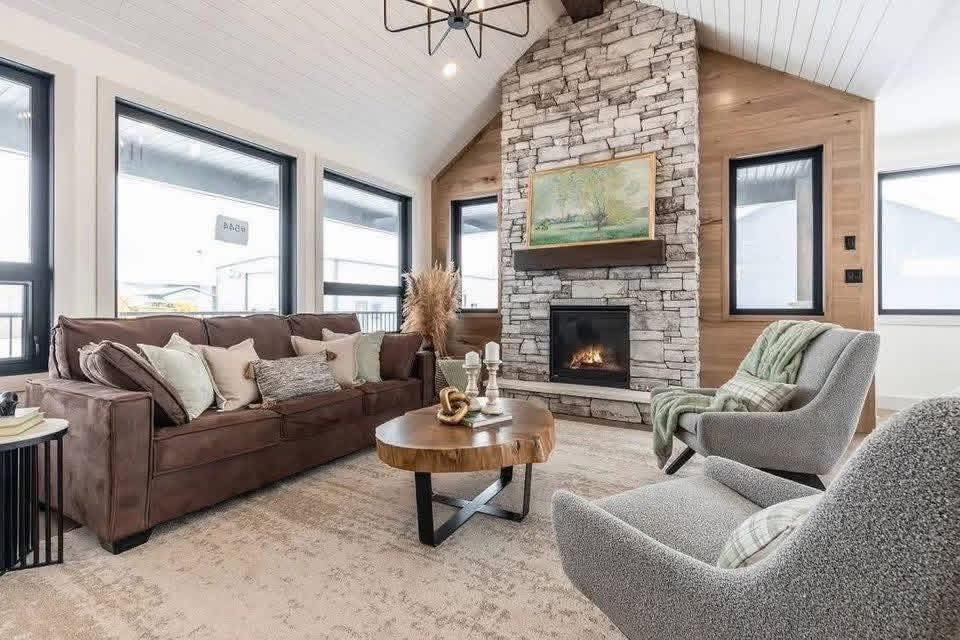 Cozy living room with a stone fireplace, brown sofa, gray armchairs, and a rustic wooden table. Large windows allow natural light, creating a warm ambiance.