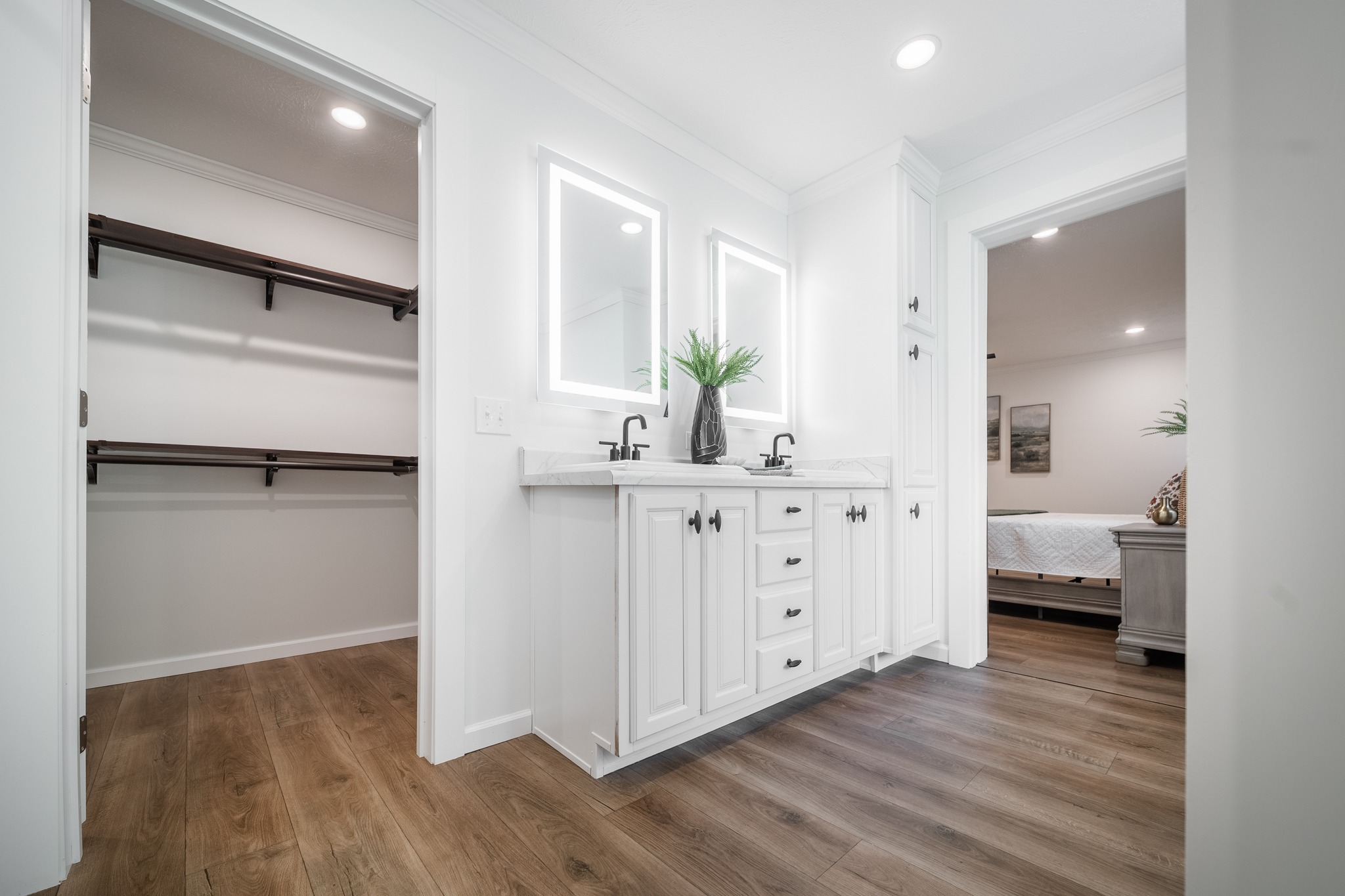 Bright bathroom with a wooden floor, featuring a white double vanity and illuminated mirrors. Open doorway to a bedroom on the right and closet on the left.