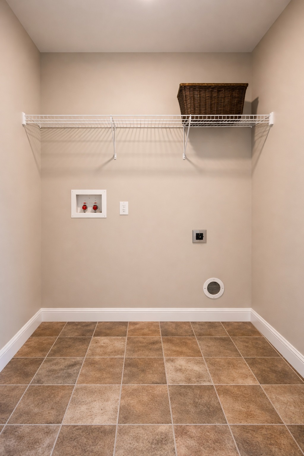 Laundry room with beige walls and tan tiled floor. A white wire shelf with a brown wicker basket is mounted on the wall above dryer and washer hookups.