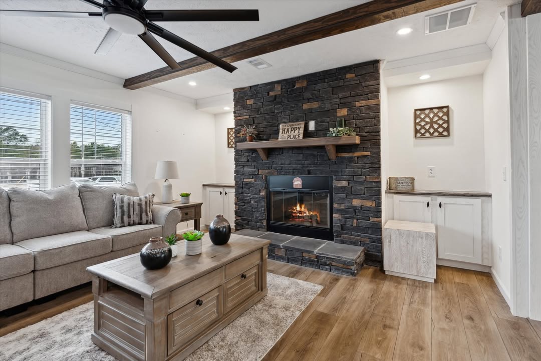Cozy inviting living room interior in modern modular home featuring rustic stone accent fireplace with electric insert and burning flames, dark wooden mantel shelf displaying farmhouse sign "The coziest happiest place" and potted succulents, exposed dark ceiling beams with recessed lighting, black industrial ceiling fan, neutral beige sectional sofa with throw pillows, distressed wooden coffee table topped with black vases and plants, light gray luxury vinyl plank flooring, large windows with white blinds letting in natural daylight, built-in white cabinets, elegant transitional farmhouse-style manufactured home great room view