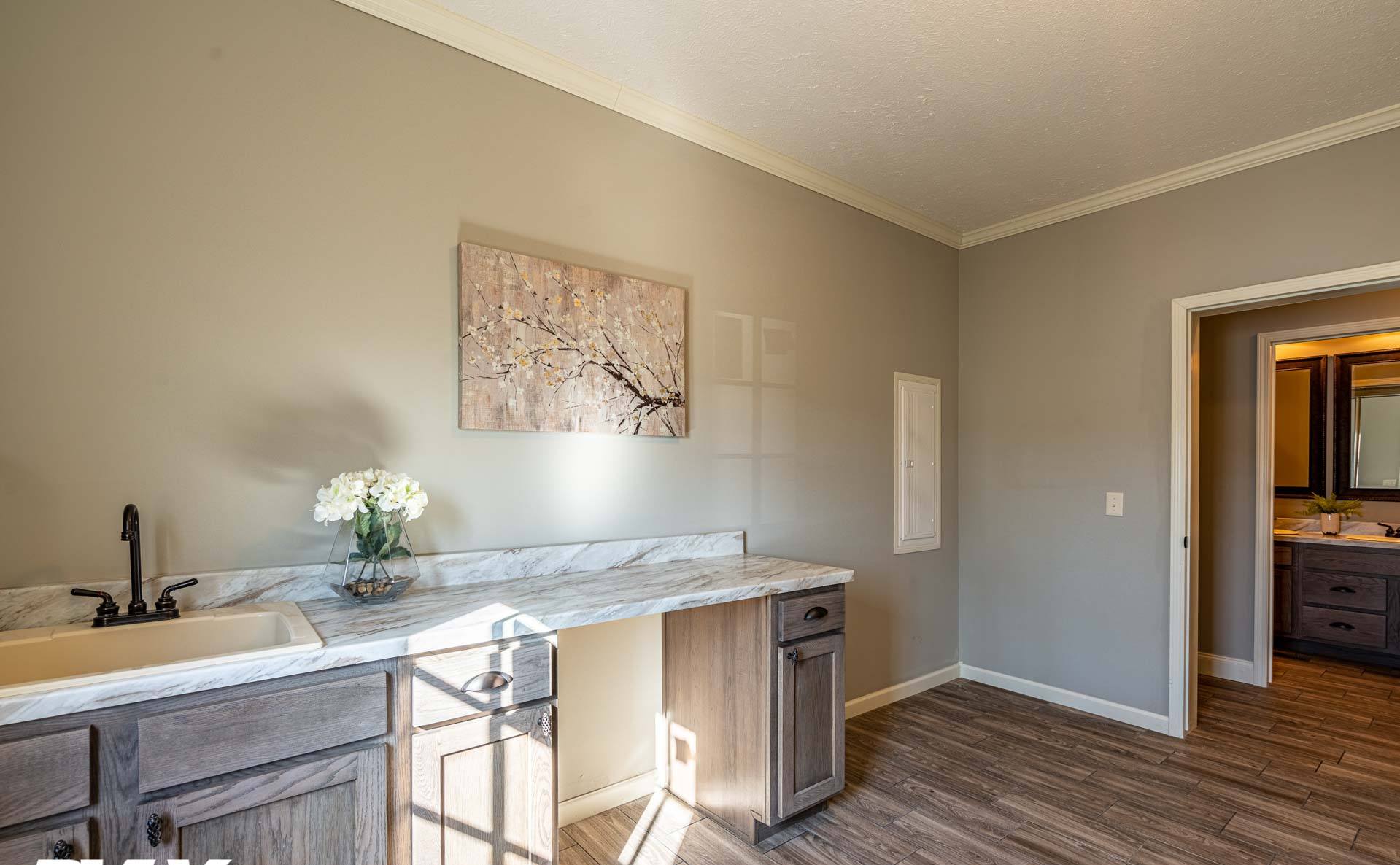 A bright, modern laundry room features a wood floor, marble countertop, and sink. A floral painting and vase with white flowers add elegance.