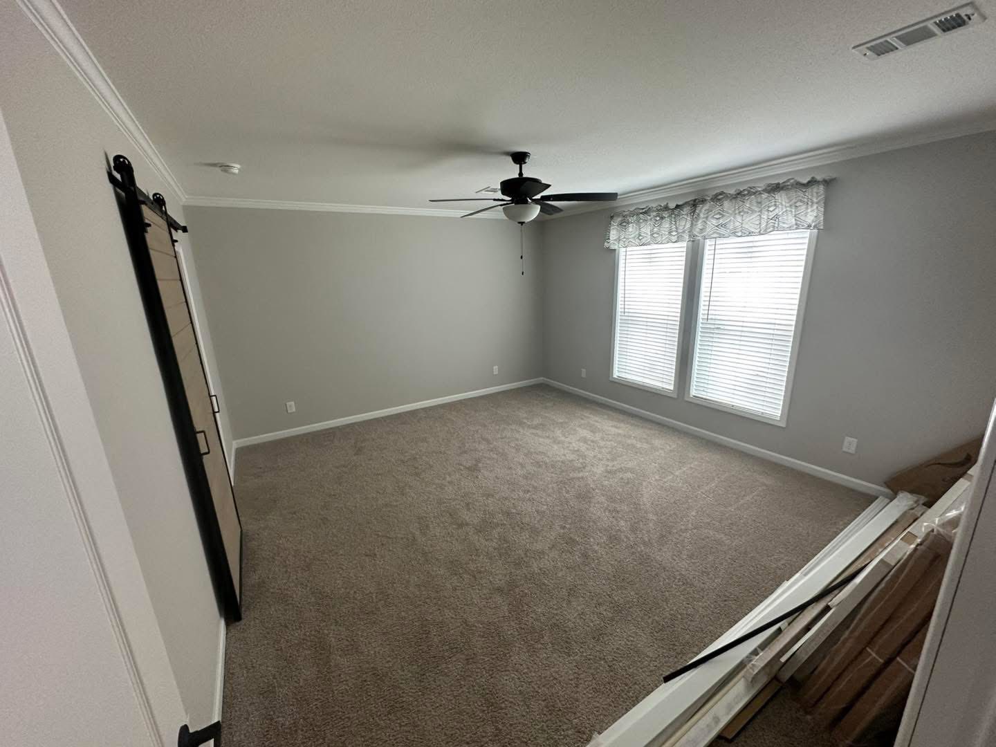 Spacious empty primary bedroom interior in modern manufactured home, light gray painted walls with white crown molding and baseboards, neutral beige carpet flooring, large double windows with white blinds and patterned valance curtains allowing natural light, black ceiling fan with light kit, sliding barn door entry with black hardware, open unfurnished layout ready for customization, bright airy master suite in 2025 modular double-wide home construction