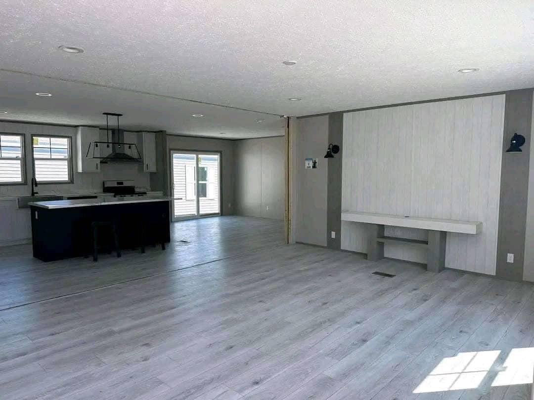 Spacious open-concept great room and kitchen interior in unfurnished modern manufactured home, vaulted textured ceiling with recessed lighting, light gray luxury vinyl plank flooring throughout, large central kitchen island with black base and white quartz countertop, white shaker cabinets, stainless steel range hood, sliding glass patio doors with blinds bringing natural light, accent shiplap wall in light wood tones with floating console shelf and black wall sconces, bright airy empty living space ready for furniture in 2025 double-wide modular home design