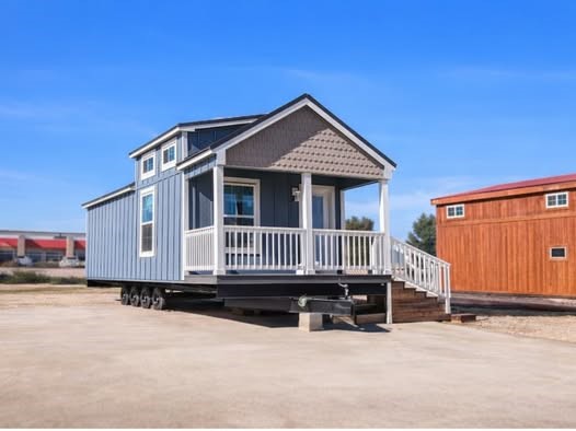 A compact, blue tiny house on wheels is parked on a concrete surface under a clear blue sky. The house has a small porch and white railing, conveying a cozy, welcoming feel.