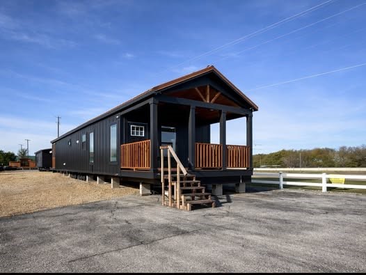 A modern black tiny house with a wooden porch sits on a gravel lot under a clear blue sky. The setting feels serene and spacious.