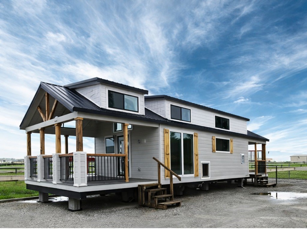 Modern light gray tiny house on wheels with a dark metal roof, a covered porch with wooden beams and railing, large windows, wooden shutters, and stairs leading to the entrance, set against a backdrop of a bright blue sky with wispy clouds.