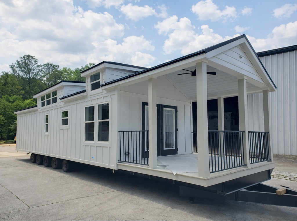 A charming white tiny home on wheels with a dark roof and black accents, featuring a welcoming covered porch with white columns and railings, multiple large windows, and dormer windows on the roof, all set against a backdrop of lush green trees and a partly cloudy sky.