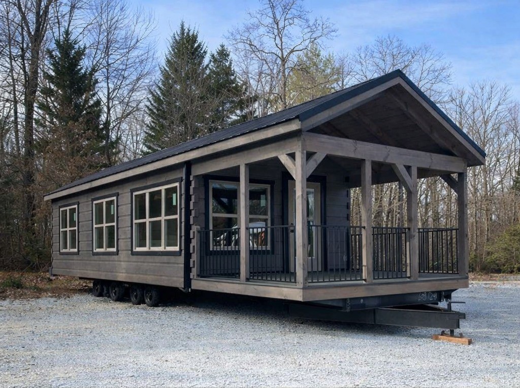 Modern dark gray tiny home on wheels with a covered porch, black metal railings, multiple large windows, and a dark roof, set against a backdrop of bare trees and a clear sky, showcasing a stylish and portable dwelling.