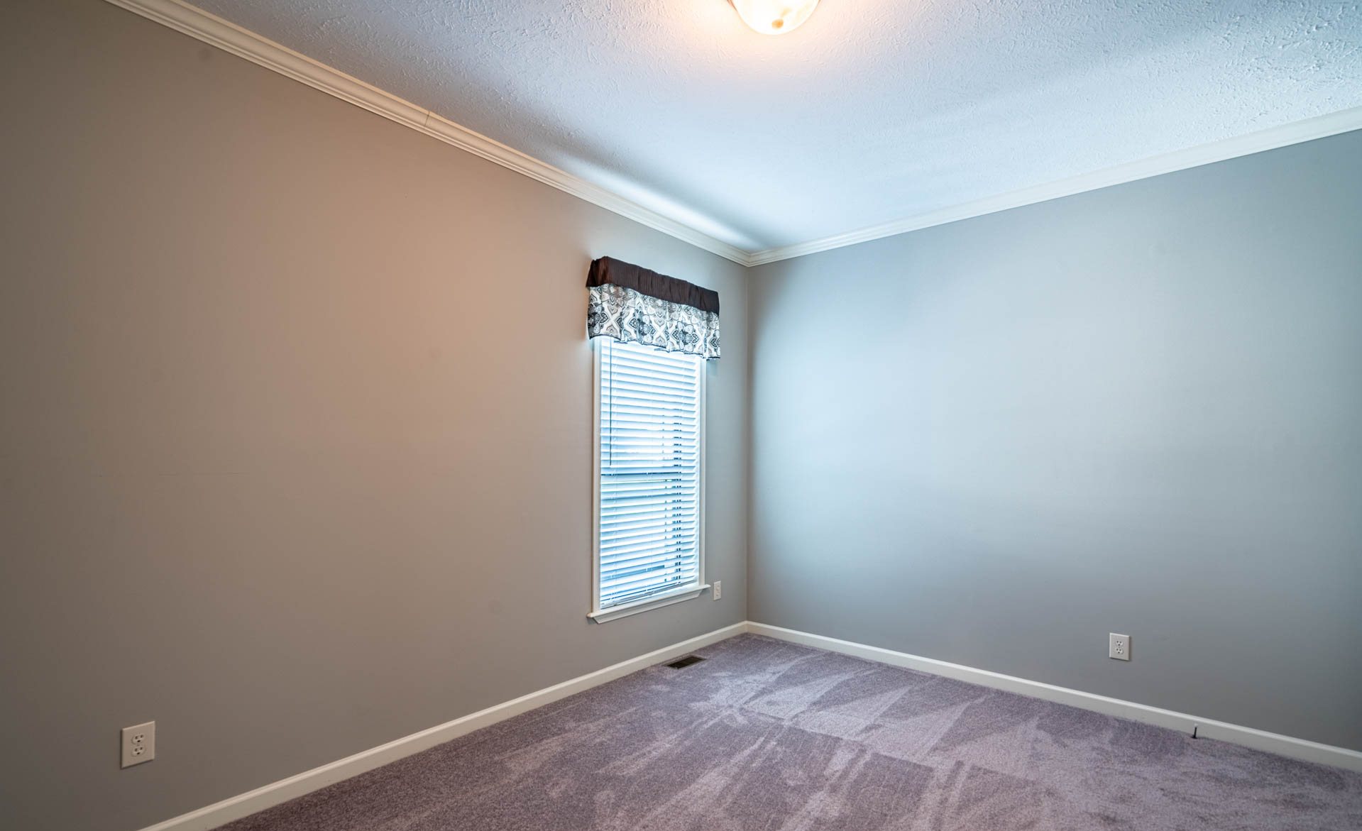 A small, empty room with light gray walls, a single window with a dark valance, and soft purple carpet. The space feels calm and clean.